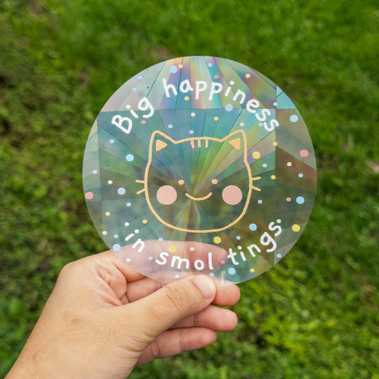 Hand holding a colorful suncatchersticker with a cat illustration and text against a grass background
