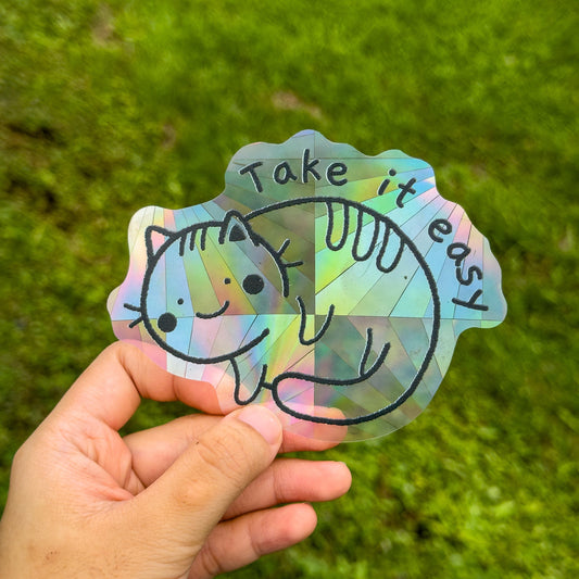 Hand holding a suncatcher sticker with a cat illustration and 'Take it easy' text against a green grass background.