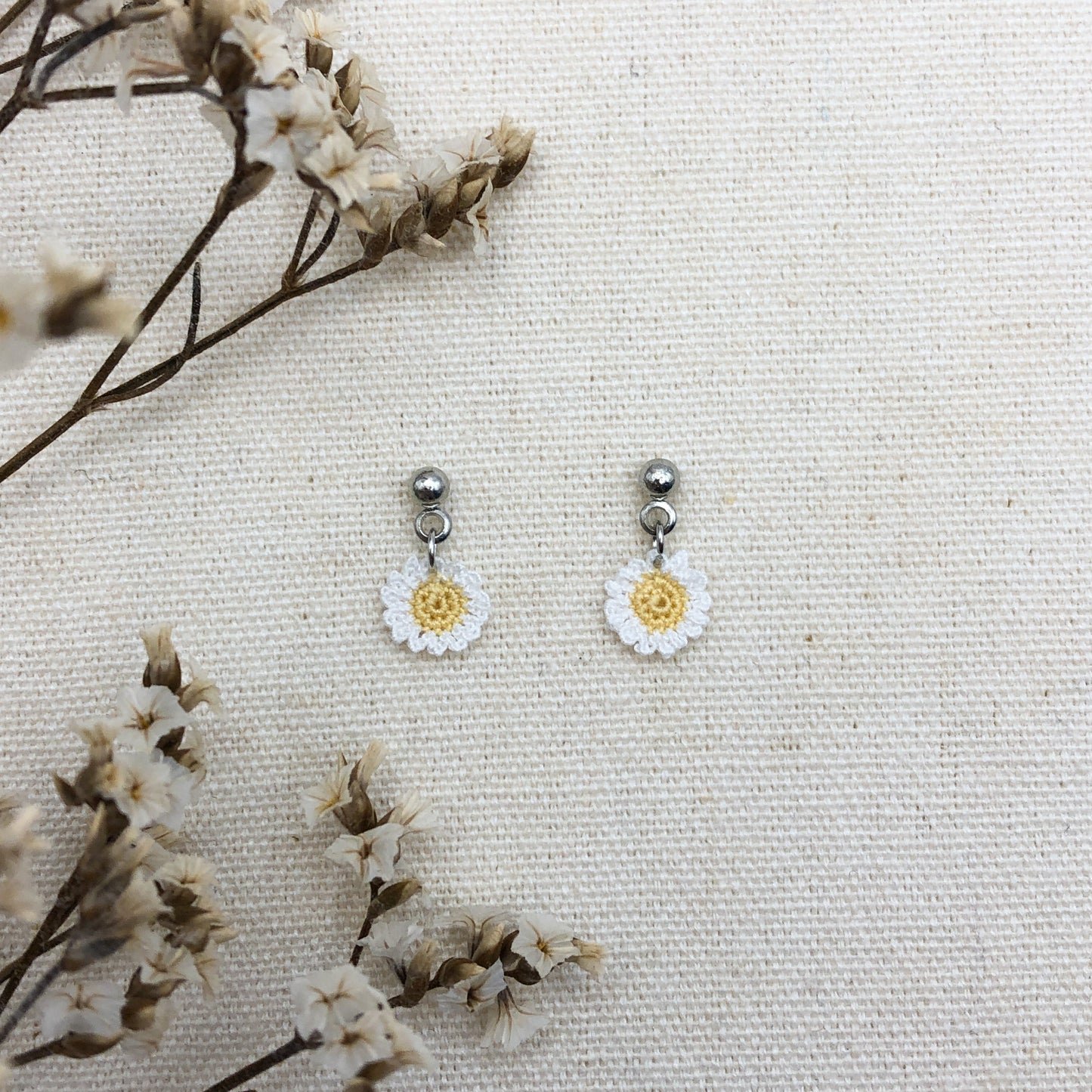 Daisy-shaped earrings on a textured white surface with dried flowers.