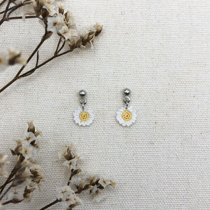 Daisy-shaped earrings on a textured white surface with dried flowers.